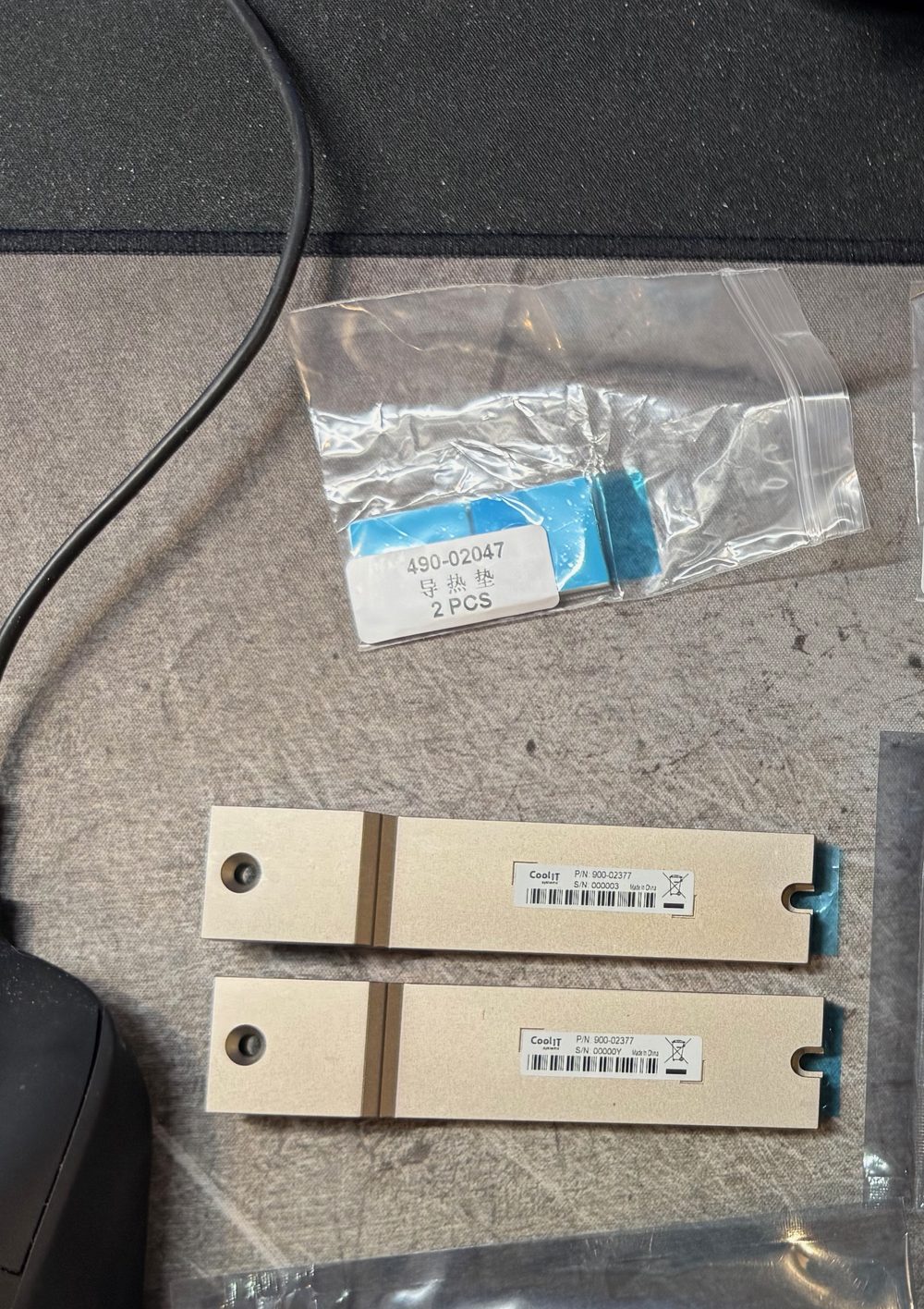 Two champagne CoolIT M.2 heatsink plates, part number 900-02377, with spare thermal pads in a bag.