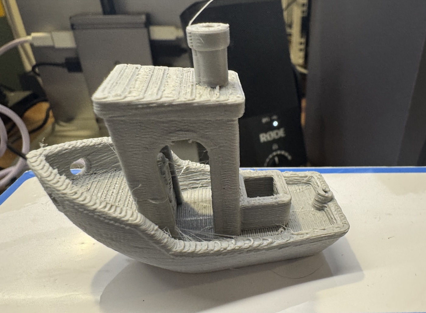 First print off the H2C: a matte grey PLA Benchy tugboat on the workbench, clean hull and chimney with faint stringing wisps.