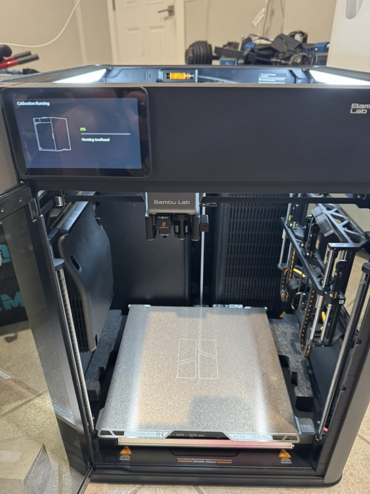 The H2C on its stand mid-first-calibration: touchscreen reads Calibration Running, Homing toolhead, with the textured build plate and dual-nozzle toolhead visible in the open chamber.
