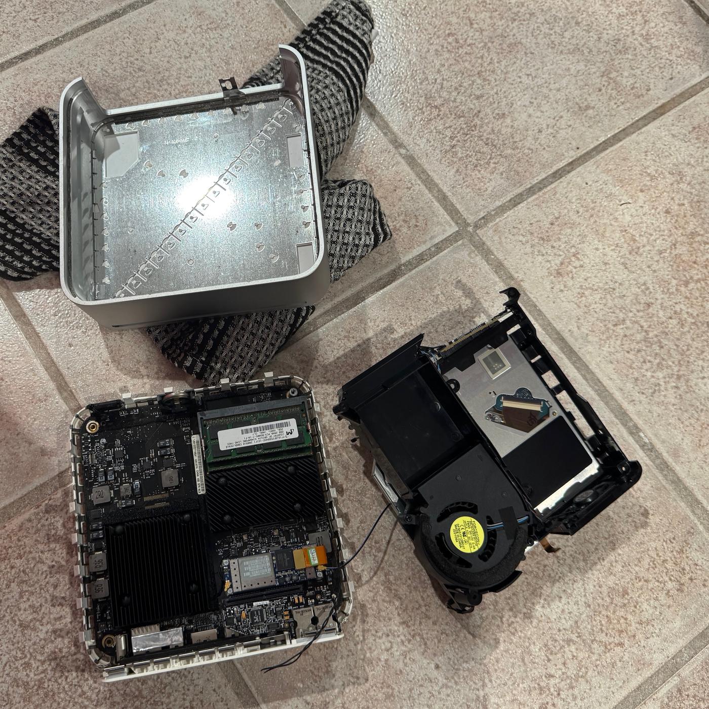Disassembled 2009 Mac Mini: empty aluminum unibody, logic board with RAM, and fan cage laid out on tile